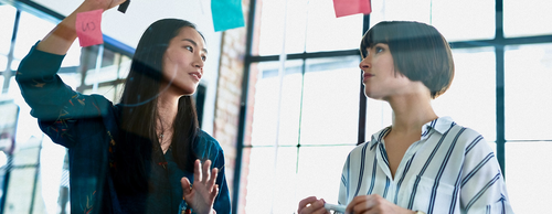 Two women collaborate in a modern office, brainstorming ideas on a glass wall covered with colorful sticky notes. One writes while the other holds a marker, engaged in discussion.