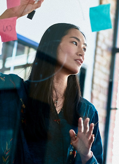 A young Asian woman in a blue floral top gazes thoughtfully at colorful sticky notes on a glass wall, her hand raised as if brainstorming or organizing ideas in a modern workspace.