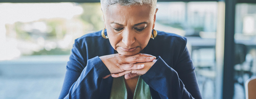 A middle-aged woman with short gray hair and gold hoop earrings sits with her chin resting on clasped hands, looking downward with a thoughtful or concerned expression, wearing a blue blazer.