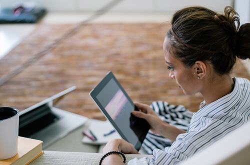 A young woman with dark hair in a bun, wearing a striped shirt, sits at a desk using a tablet. A laptop, coffee mug, and book are nearby. She smiles while browsing, in a bright, modern space.
