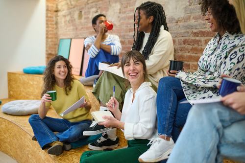 A diverse group of young people sitting casually on wooden steps, laughing and chatting. They hold notebooks and drinks in a creative space with brick walls, suggesting a relaxed brainstorming or study session.
