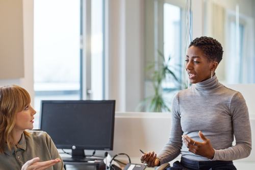 Two women having a conversation in a modern office. One sits at a desk gesturing, while the other perches on the desk's edge, smiling. A computer monitor and desk phone are visible in the bright, open workspace.