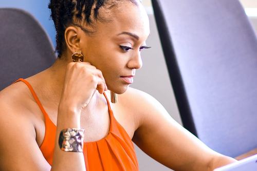 A focused woman with twisted updo hair, wearing an orange top and silver bracelet, sits at a desk working on a laptop, holding a pen thoughtfully to her chin.