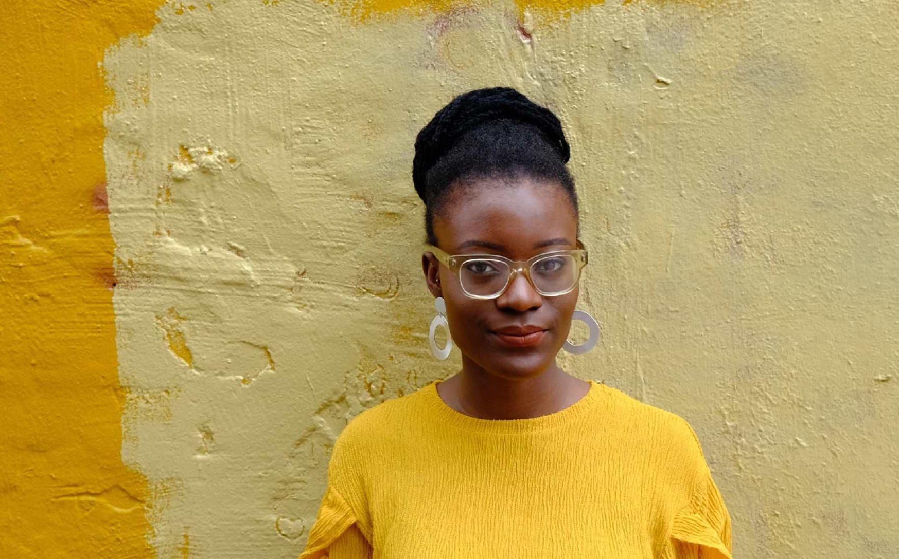 A young woman wearing a yellow top, clear-framed glasses, and hoop earrings, with her natural hair in an updo, poses against a textured yellow wall.