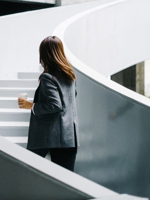 A woman with long brown hair, wearing a gray blazer and black pants, holds a coffee cup while standing on a curved white staircase, viewed from behind in a modern, minimalist setting.