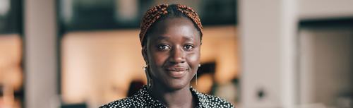 A young woman with braided hair pinned up, wearing dangling earrings and a black polka-dot top, smiles softly at the camera in a modern indoor setting with blurred background.