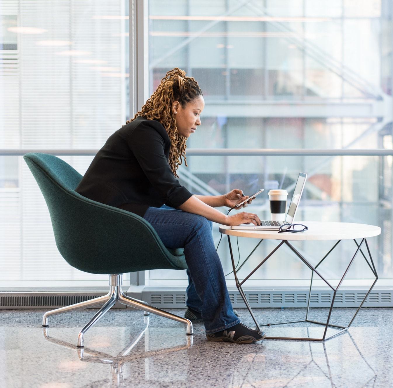 A woman with locs sits in a teal chair by large windows, working on a laptop while holding a smartphone. A coffee cup and glasses rest on a modern geometric table beside her.