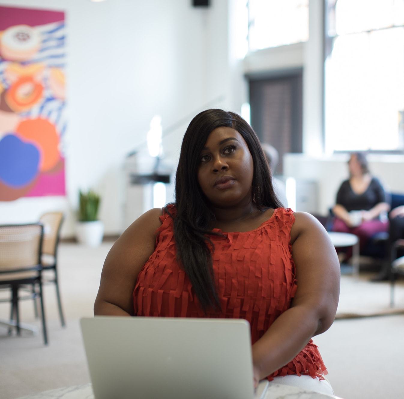 A woman in a red sleeveless top sits with a laptop in a bright, modern workspace, looking thoughtfully upward. A colorful abstract artwork hangs on the wall behind her, with another person seated in the background.