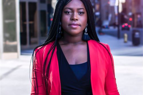 A confident young woman with long black braids and dangling earrings, wearing a bold red blazer over a black top, stands on a city street with blurred urban buildings in the background.