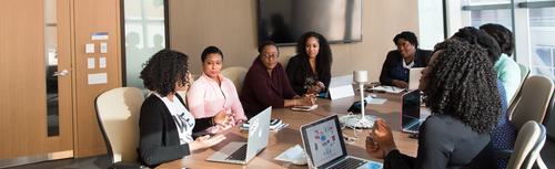 A group of professional women gathered around a conference table with laptops, engaged in a business meeting in a modern office setting with large windows.
